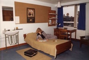Female student in 1970s suit reading textbook on a bed in Mason Hall. The room includes a sink, mirror, pinboard, desk and shelves